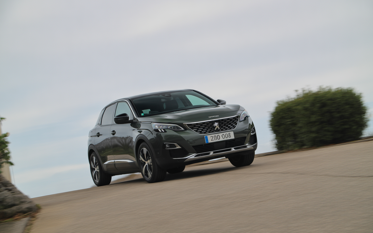 Voiture Peugeot gris foncé, en mouvement sur une route sinueuse, avec ciel nuageux en arrière-plan et verdure sur le côté.
