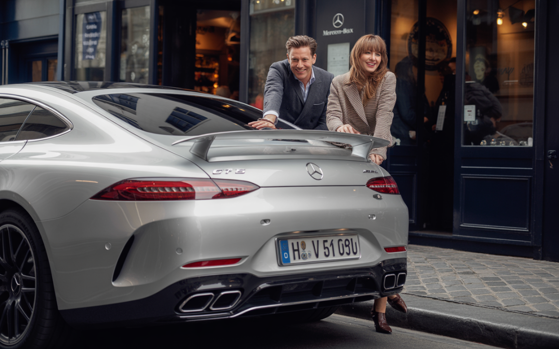 Un couple souriant pose devant une Mercedes-Benz AMG GT, garée sur une rue pavée près d'une boutique.