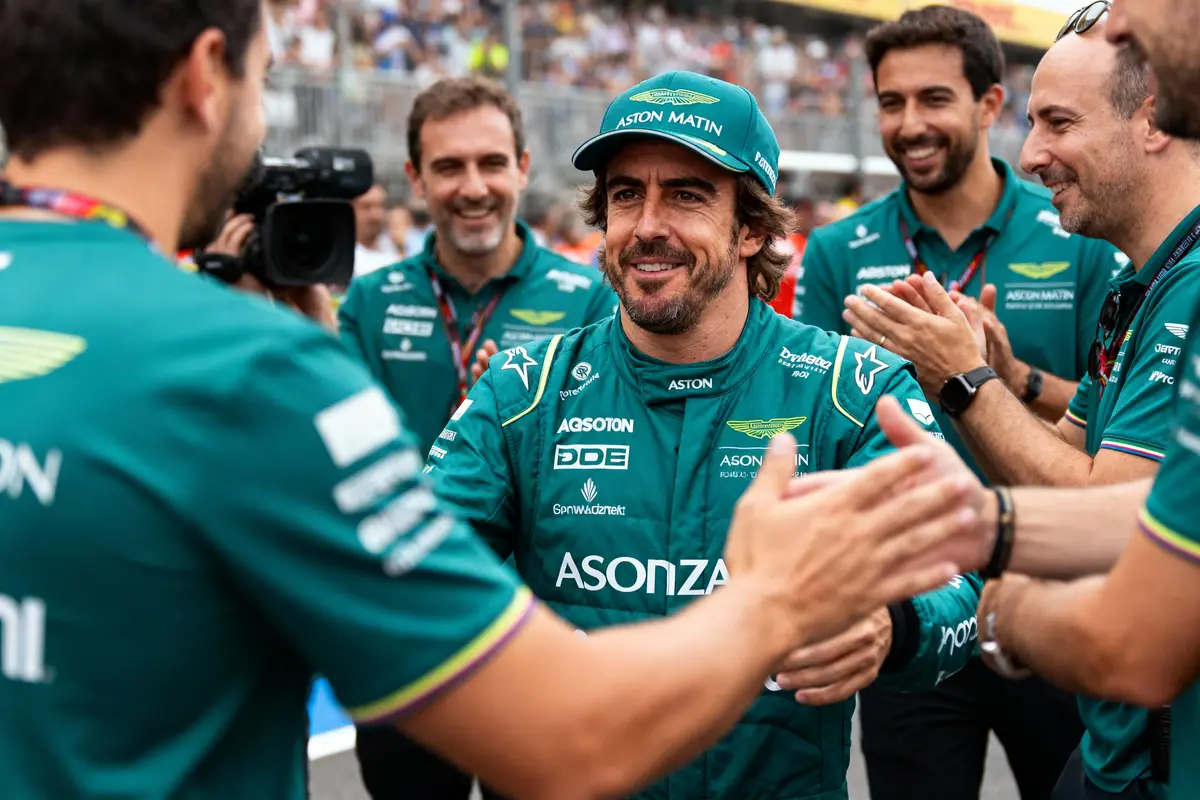 Pilote en uniforme Aston Martin, entouré de l'équipe, applaudissements et félicitations sur circuit rempli de spectateurs.