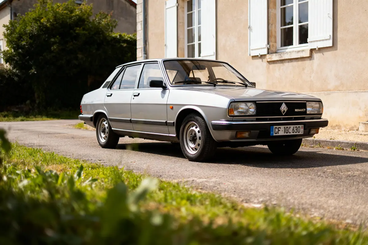 Voiture classique argentée stationnée devant une maison beige avec volets blancs, en journée ensoleillée.