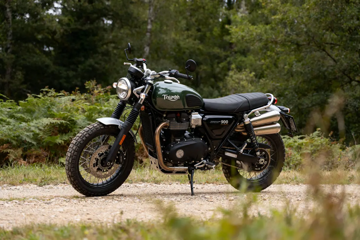 Moto rétro verte Triumph Scrambler 900 stationnée sur chemin forestier, entourée de végétation dense.