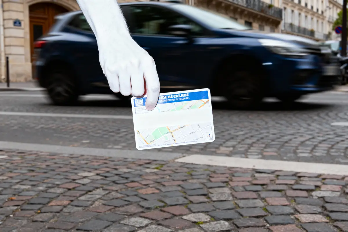 Main blanche tenant une carte devant une voiture bleue en mouvement, sur une rue pavée.