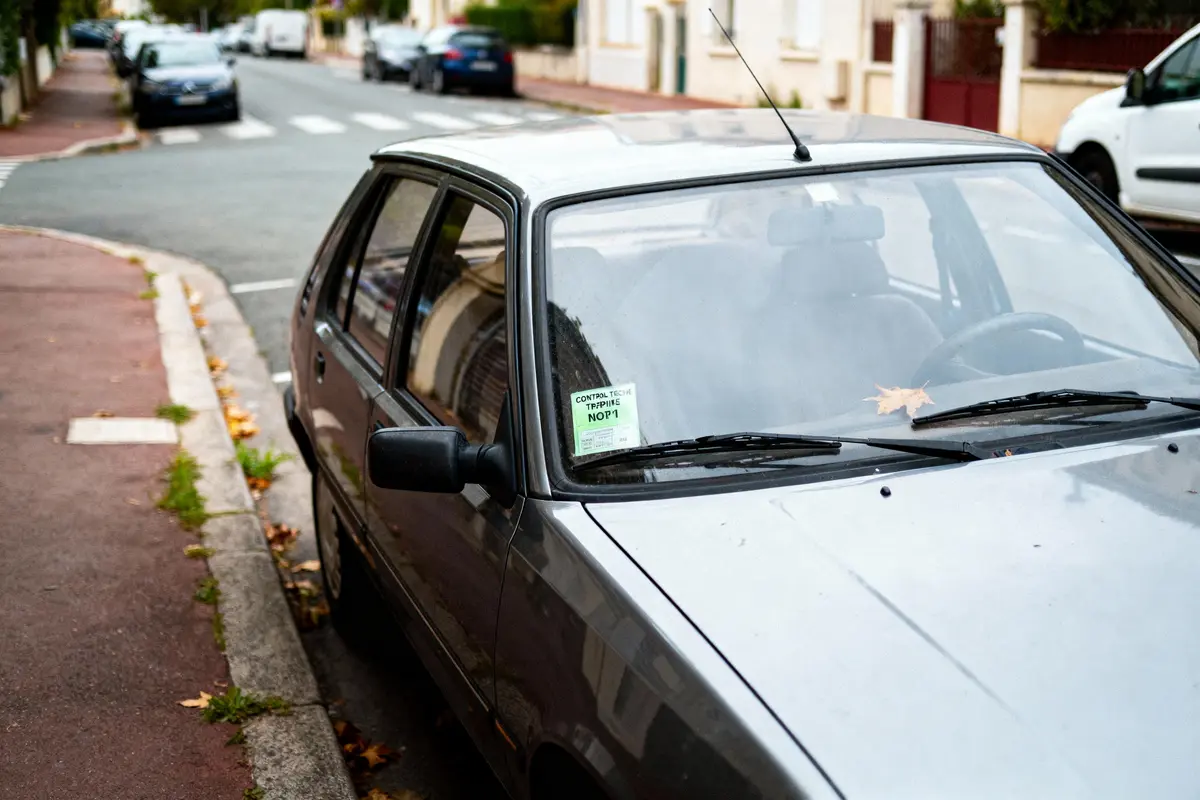 Voiture argentée garée sur rue résidentielle, feuille sur pare-brise, vignette de contrôle technique visible à l'intérieur.