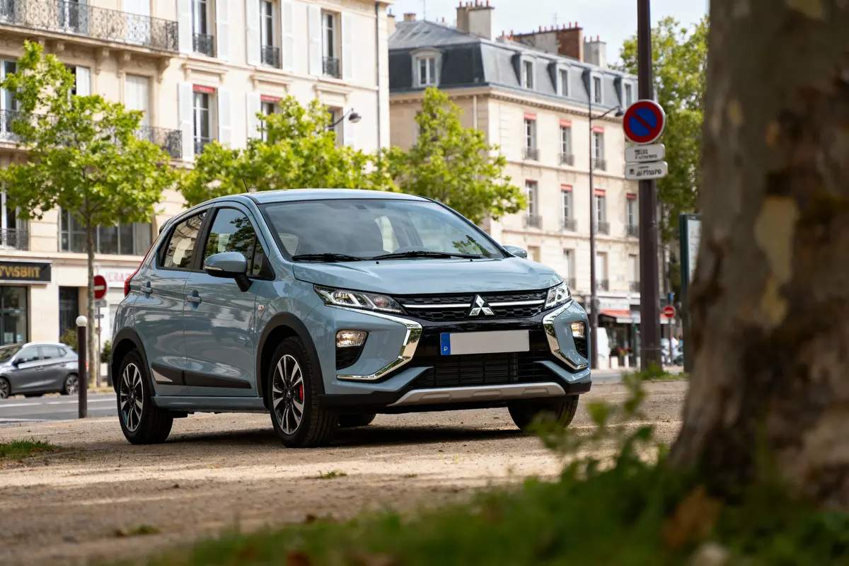 Voiture Mitsubishi grise garée en ville, devant immeubles classiques et arbres verts. Panneau stationnement interdit visible.