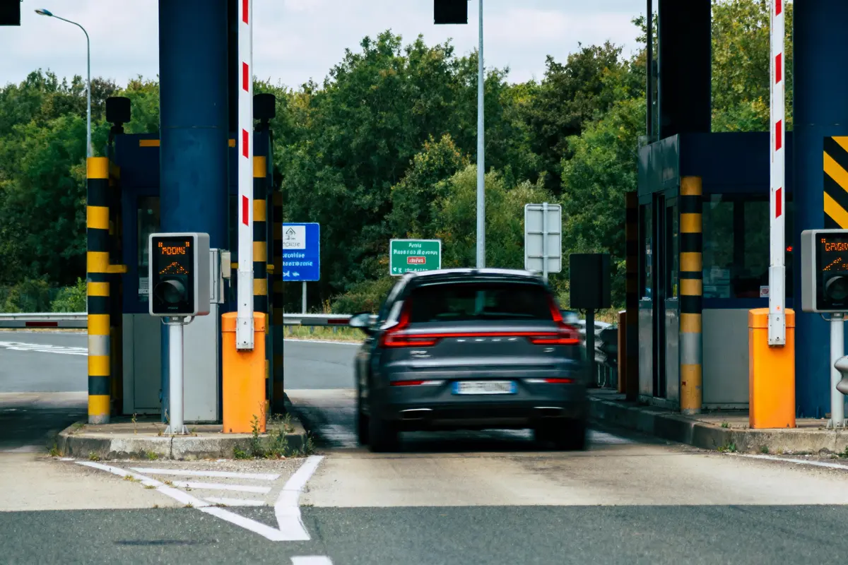 Voiture passant un péage sur une autoroute, enseignes de signalisation, barrières levées, arrière-plan forestier.