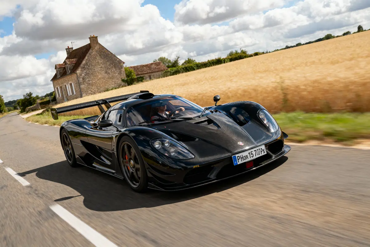 Voiture de sport noire rapide sur route de campagne, sous un ciel partiellement nuageux près d'une maison en pierre.