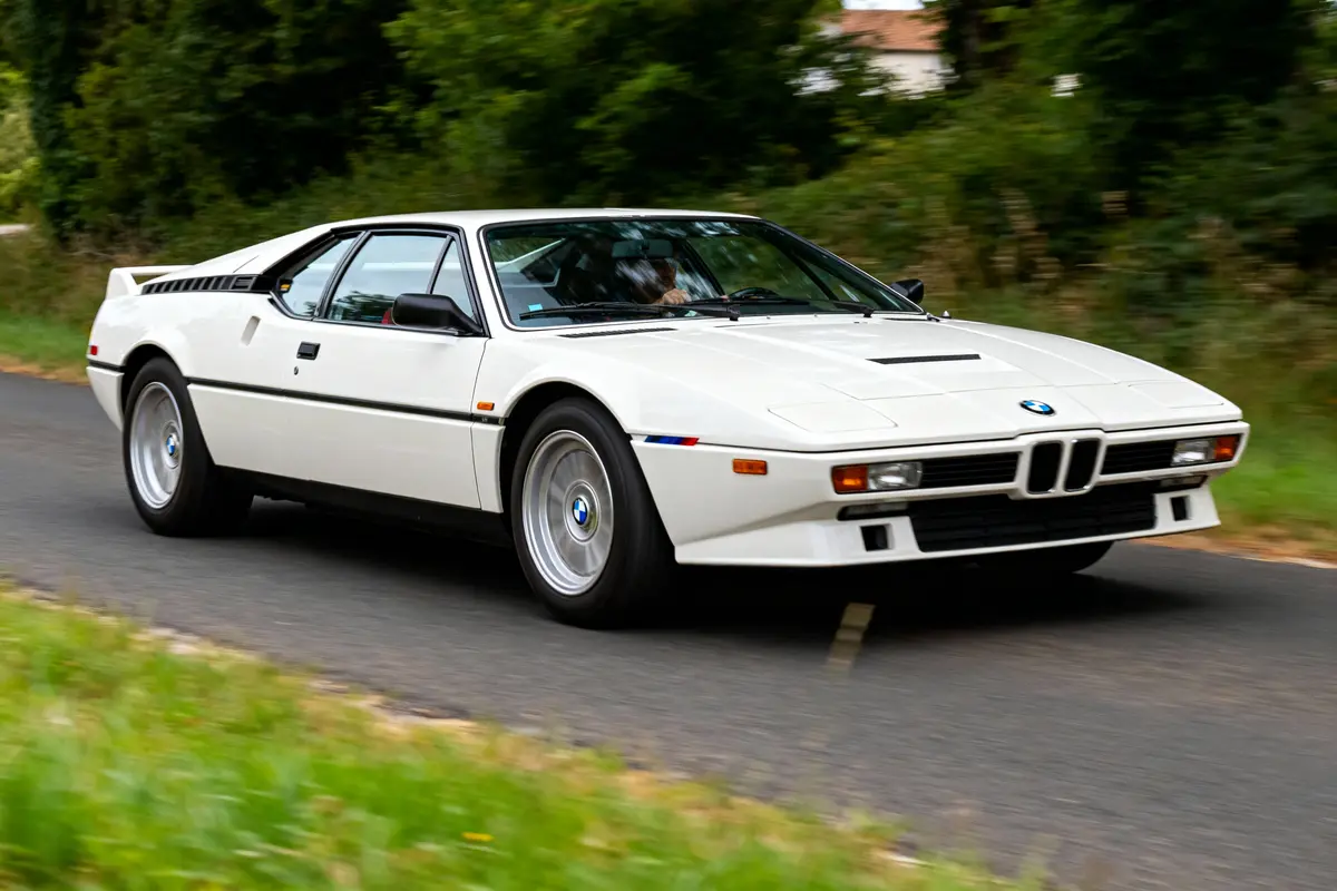 Voiture de sport blanche sur route, entourée de verdure, en mouvement, avec logo BMW sur le capot.