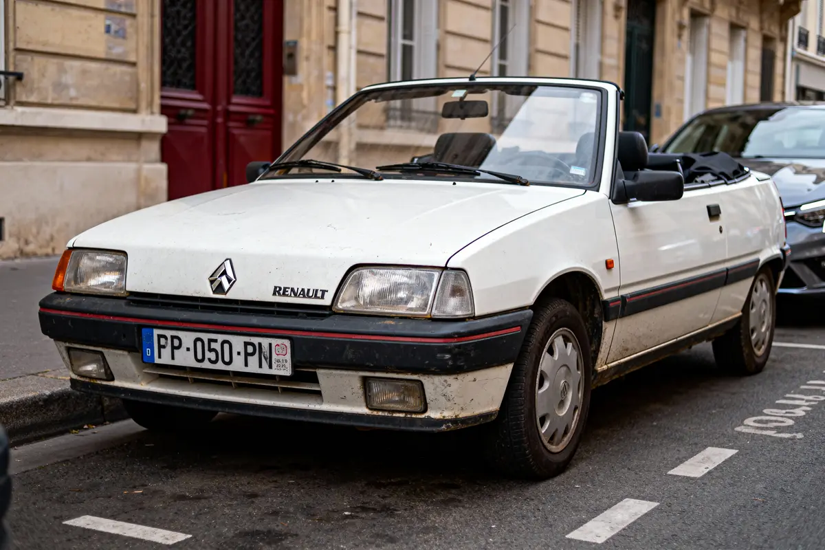 Voiture décapotable blanche Renault garée dans une rue, devant une porte rouge, bâtiments en arrière-plan.