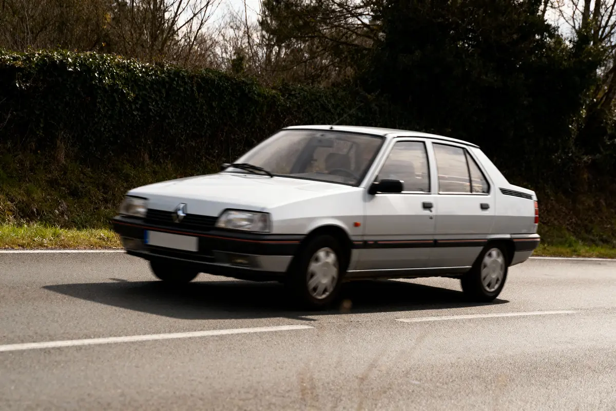 Voiture blanche en mouvement sur route, entourée d'arbres et de végétation. Journée légèrement nuageuse.