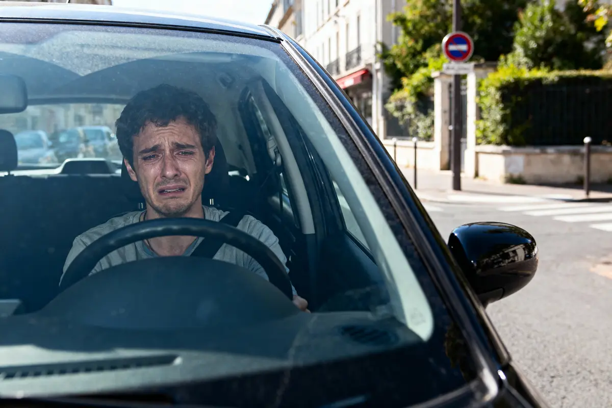 Homme pleurant au volant d'une voiture, rue ensoleillée en arrière-plan, panneau de signalisation visible.