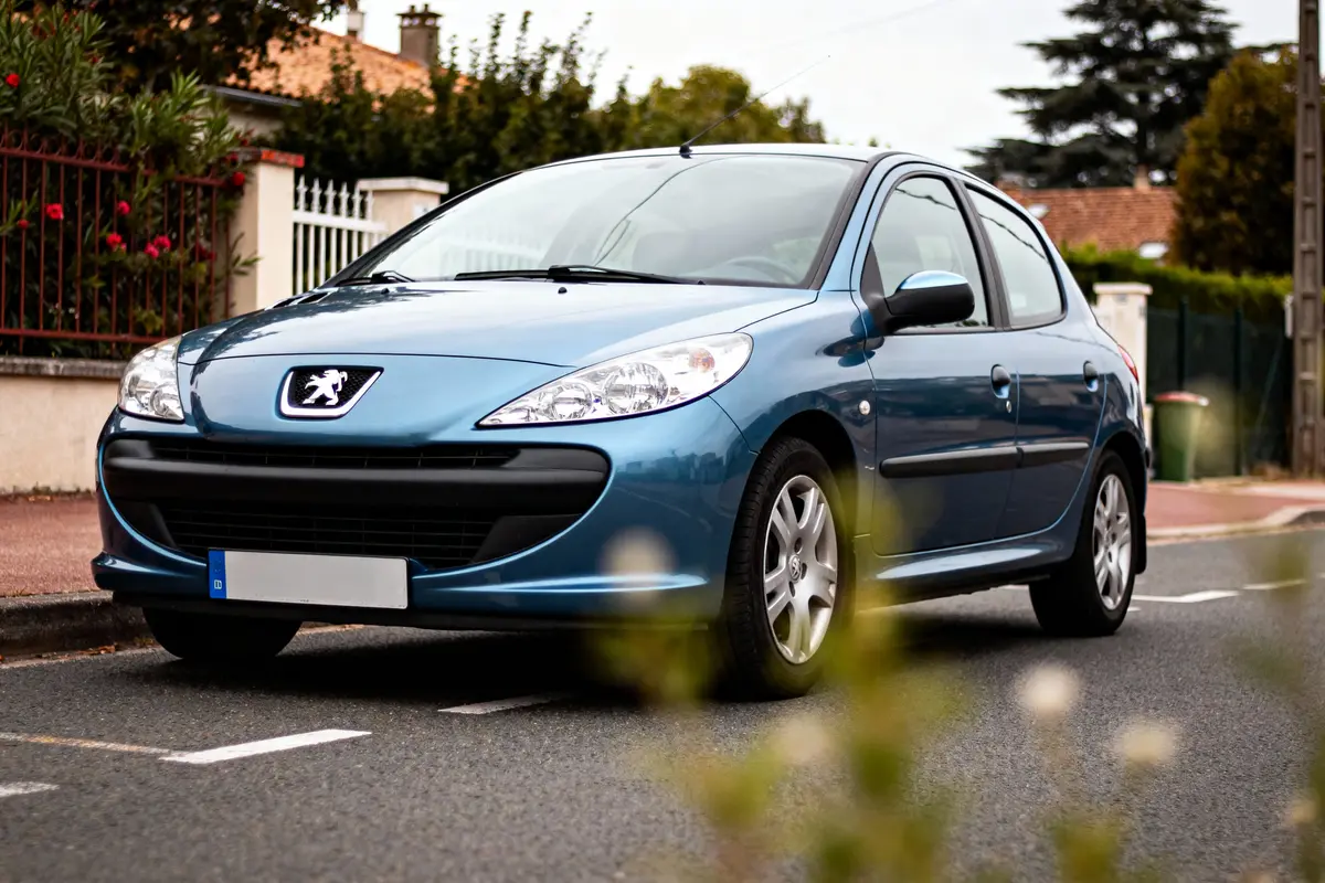 Voiture bleue Peugeot stationnée près d'une clôture, avec arrière-plan de végétation et maisons.