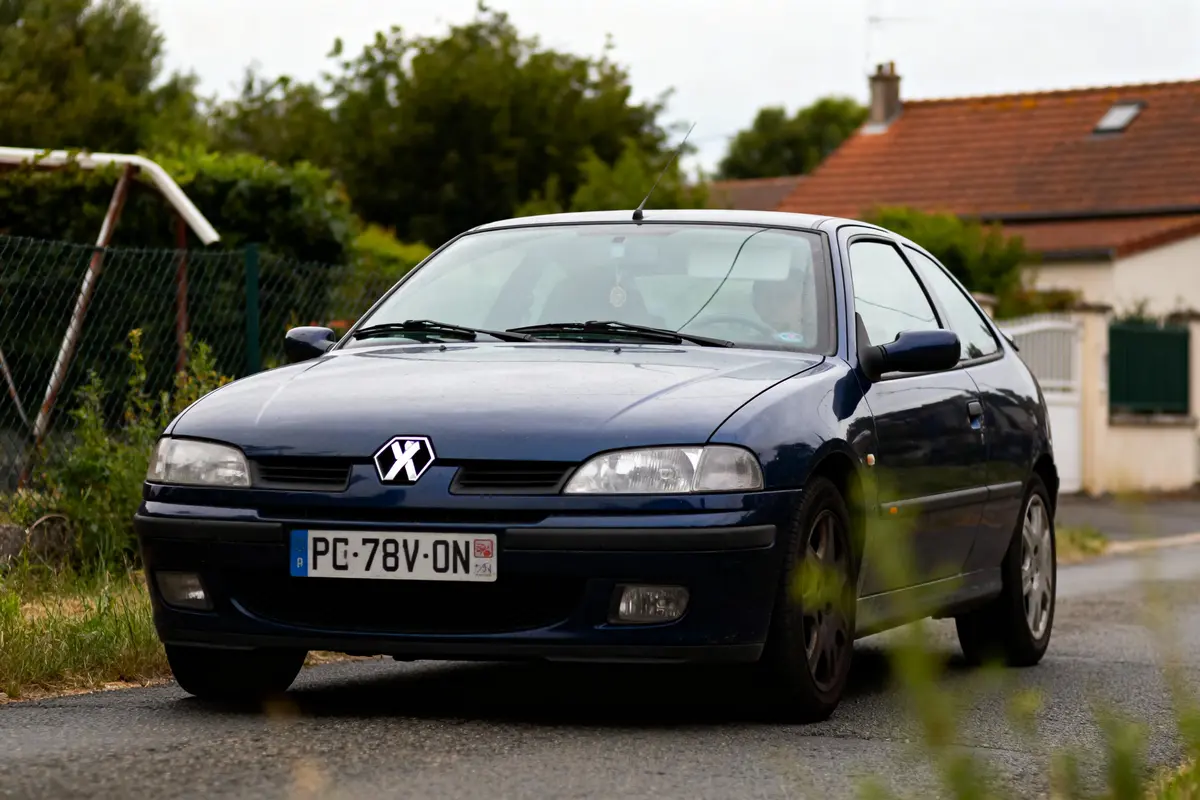 Voiture bleue garée sur la route, devant une maison avec toiture rouge et végétation environnante.