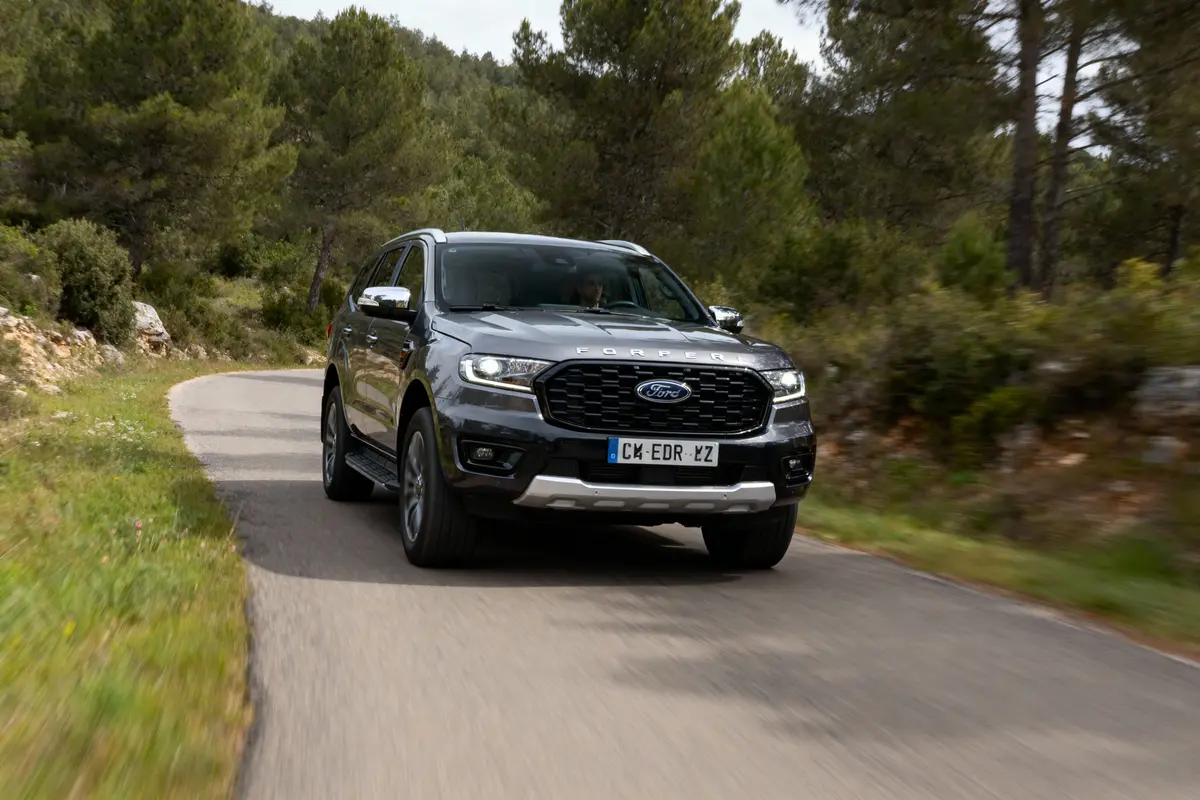 SUV Ford gris roulant sur une route de campagne bordée d'arbres et buissons, ciel légèrement nuageux.
