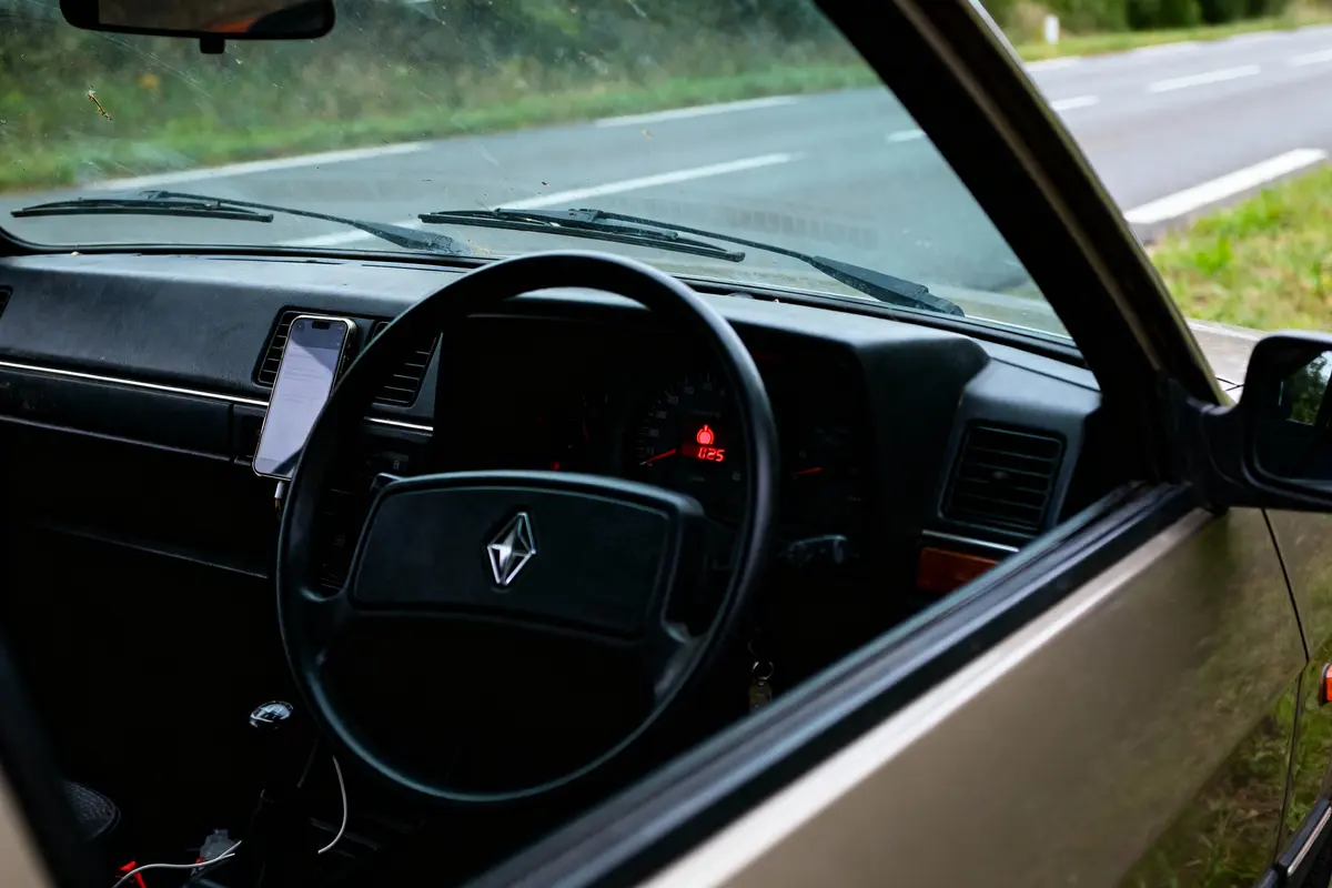 Intérieur de voiture avec volant, tableau de bord, téléphone fixé, route visible par la fenêtre, ciel partiellement nuageux.