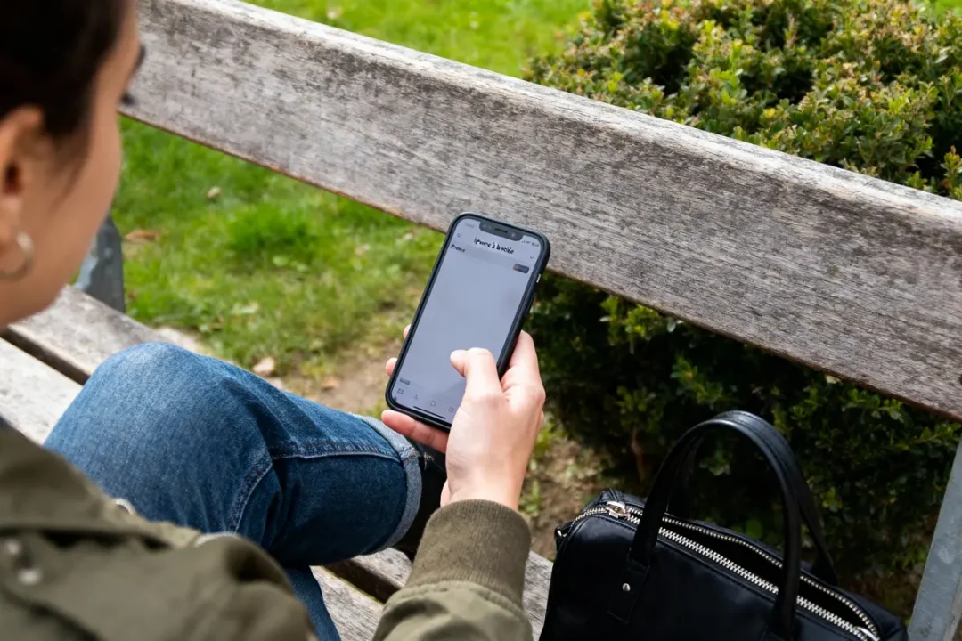 Personne sur un banc, utilisant un smartphone, veste verte, jean, sac noir à côté, parc en arrière-plan.