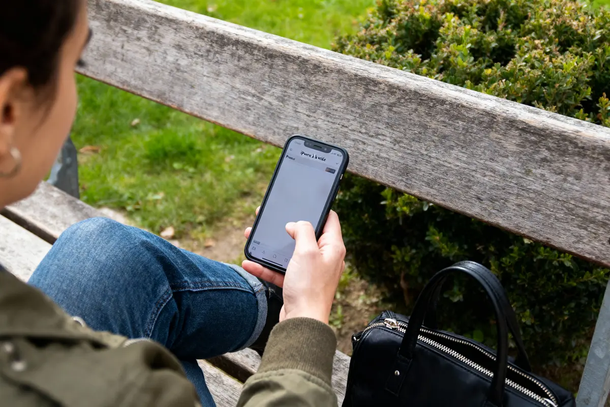 Personne sur un banc, utilisant un smartphone, veste verte, jean, sac noir à côté, parc en arrière-plan.
