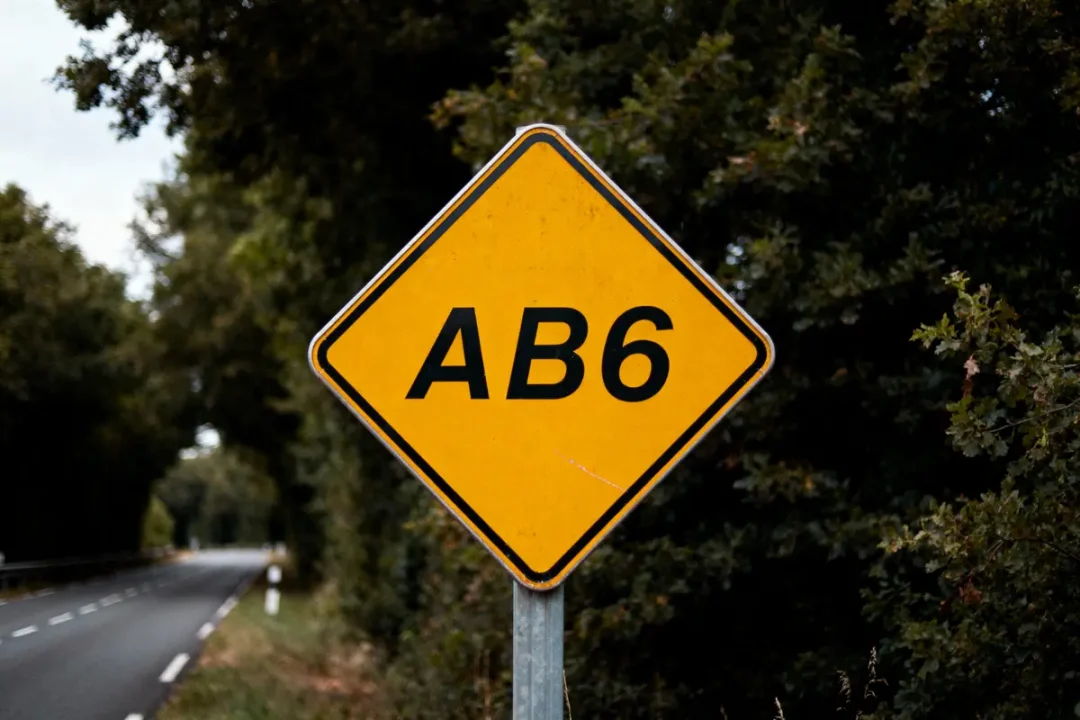Panneau routier jaune marqué "AB6", sur fond de route et arbres verts.