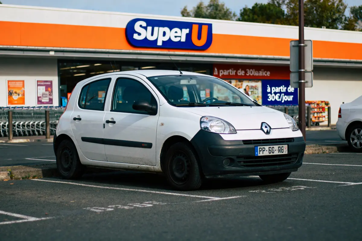Voiture blanche Renault stationnée devant un supermarché Super U, avec caddies visibles à l'arrière.