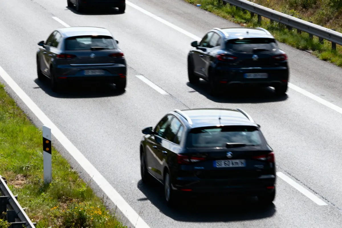 Trois voitures noires roulant sur une autoroute à deux voies, bordée de verdure et de barrières de sécurité.