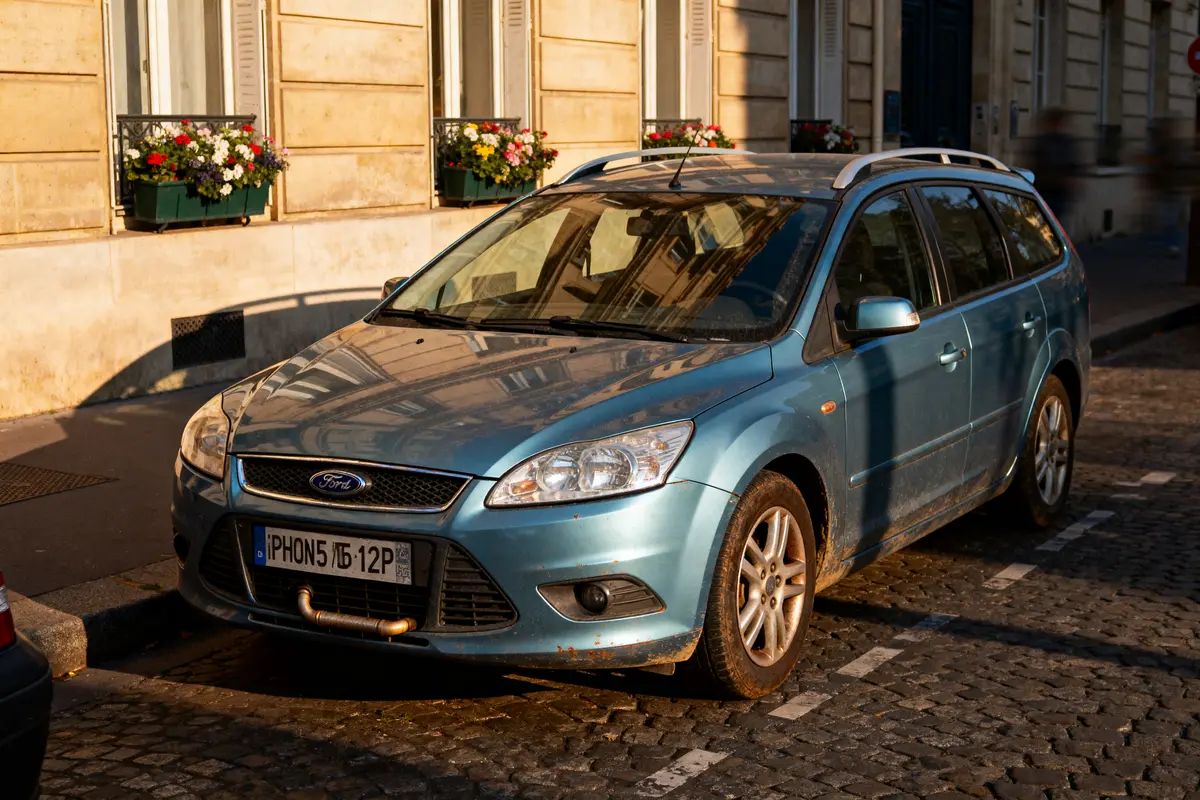 Voiture Ford bleue stationnée sur une rue pavée, bâtiments en arrière-plan avec jardinières fleuries.