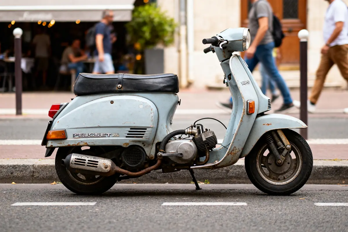Vieux scooter Peugeot bleu garé en ville, piétons flous en arrière-plan, café en terrasse derrière.