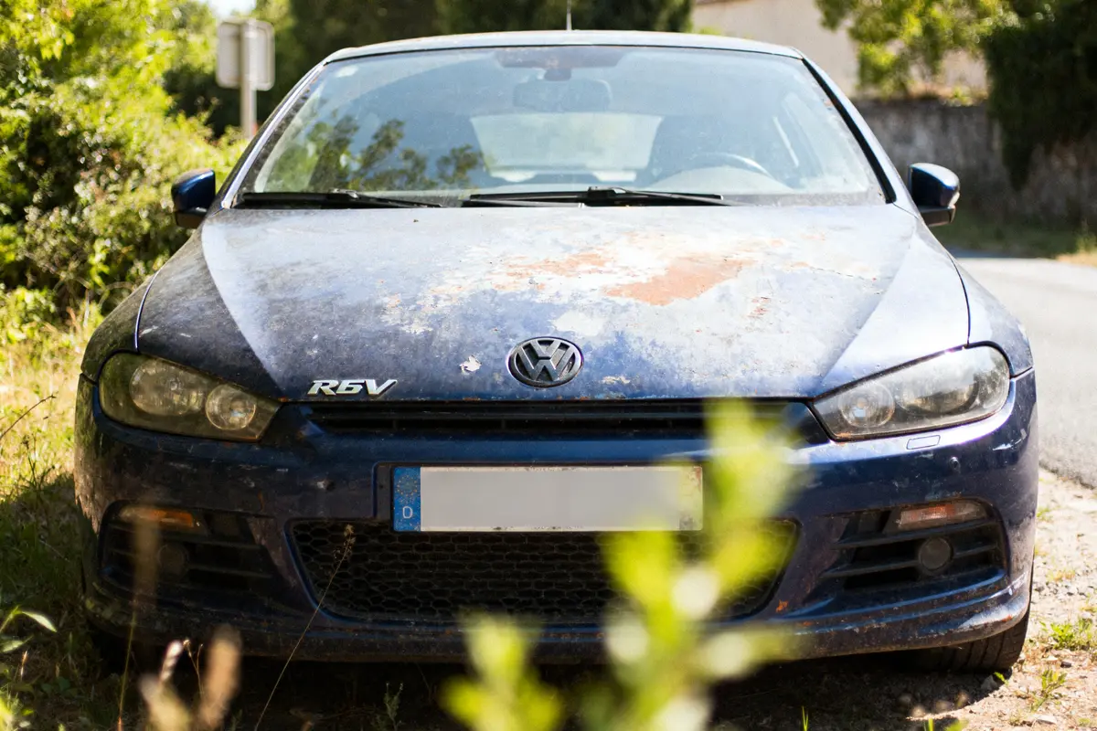 Voiture bleue usée et rouillée, garée près d'un chemin entouré de végétation.