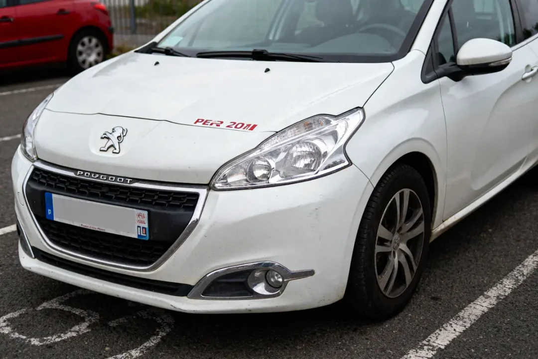 Voiture Peugeot blanche, texte "PER 20M1" sur le capot, stationnée sur asphalte. Plaque d'immatriculation floutée.