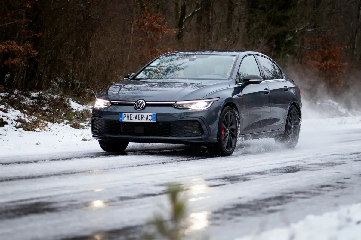 Voiture Volkswagen roulant sur route enneigée, entourée d'arbres, avec traces de neige sur la chaussée.