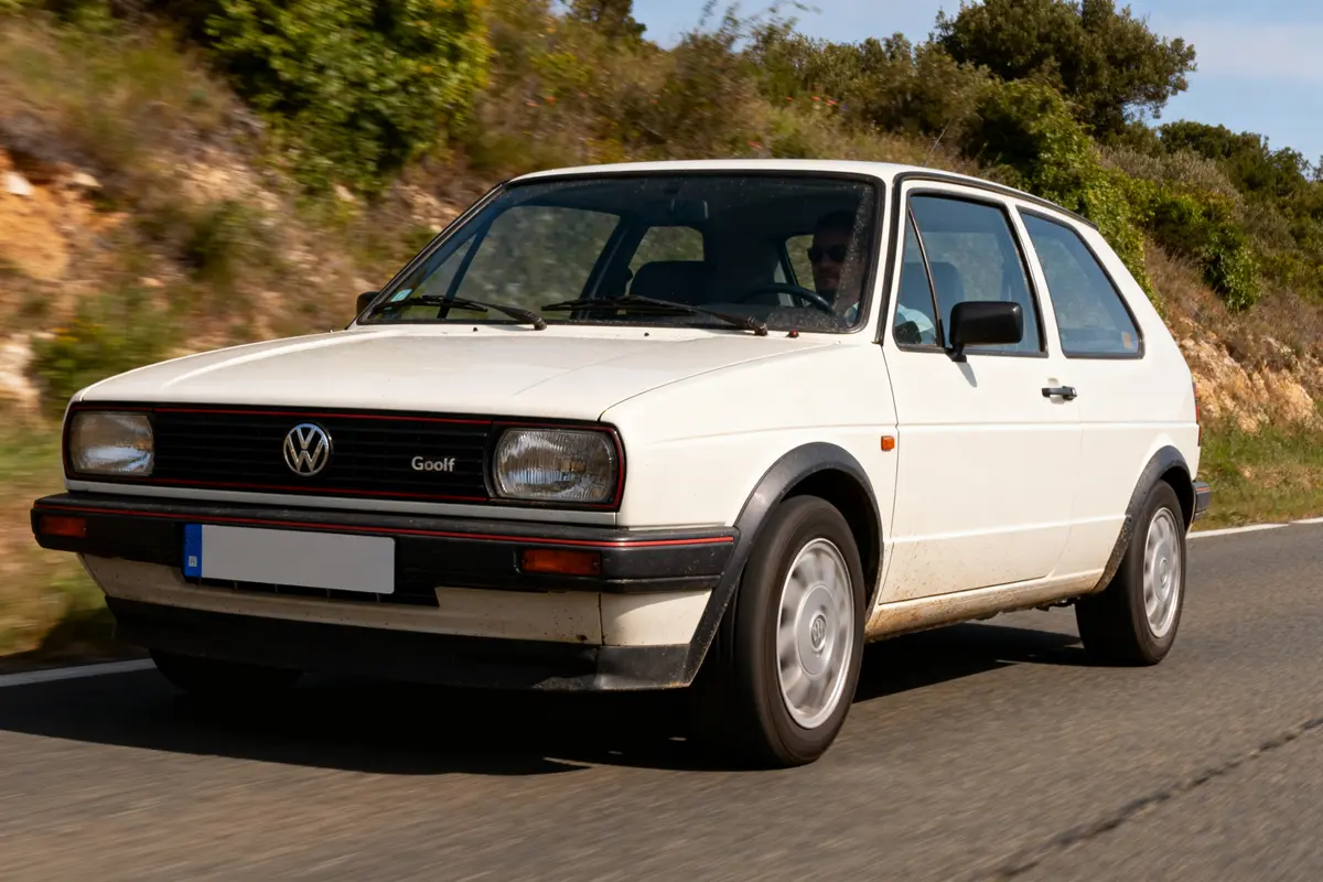 Voiture blanche Volkswagen Golf 1 1 1 roule sur une route bordée d'arbres.