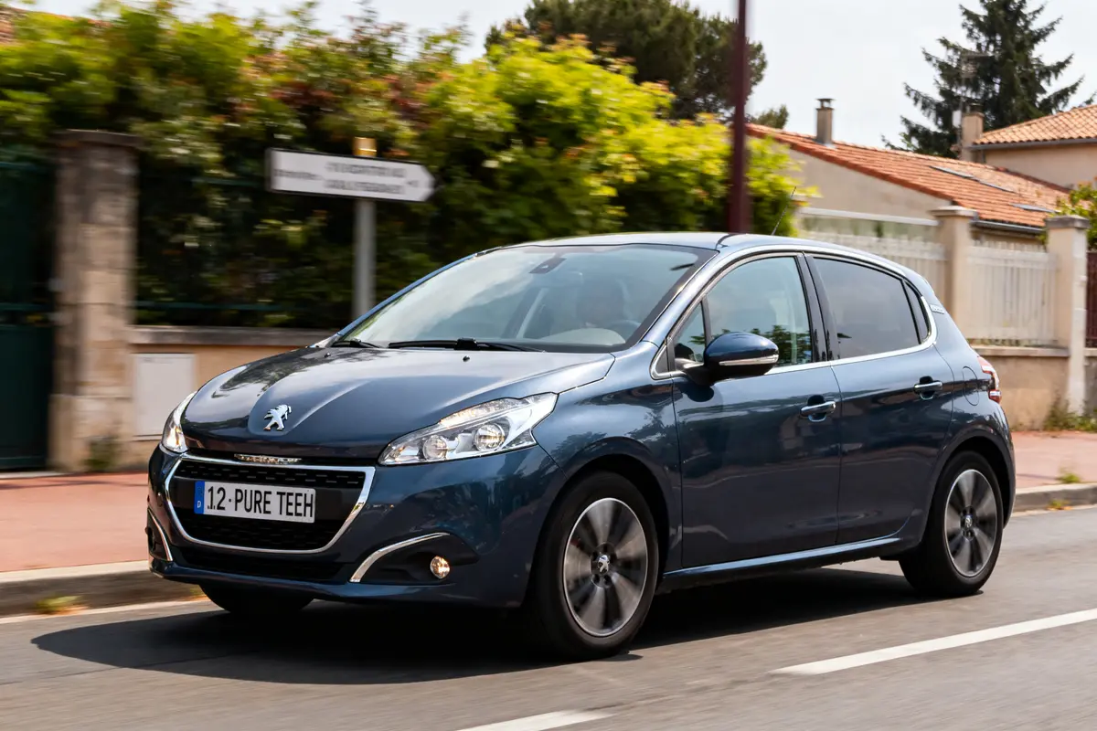 Voiture Peugeot bleue roulant dans une rue, entourée de végétation et habitations faiblement visibles.