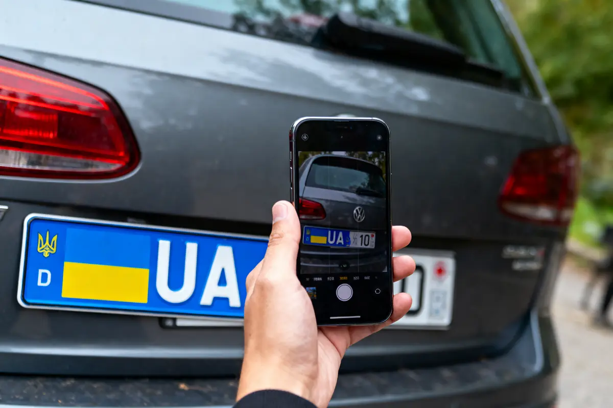 Main tenant un smartphone photographiant la plaque d'immatriculation ukrainienne d'une voiture noire stationnée.