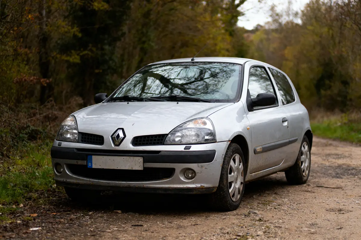 Petite voiture argentée Renault stationnée sur un chemin de terre, entourée d'arbres.